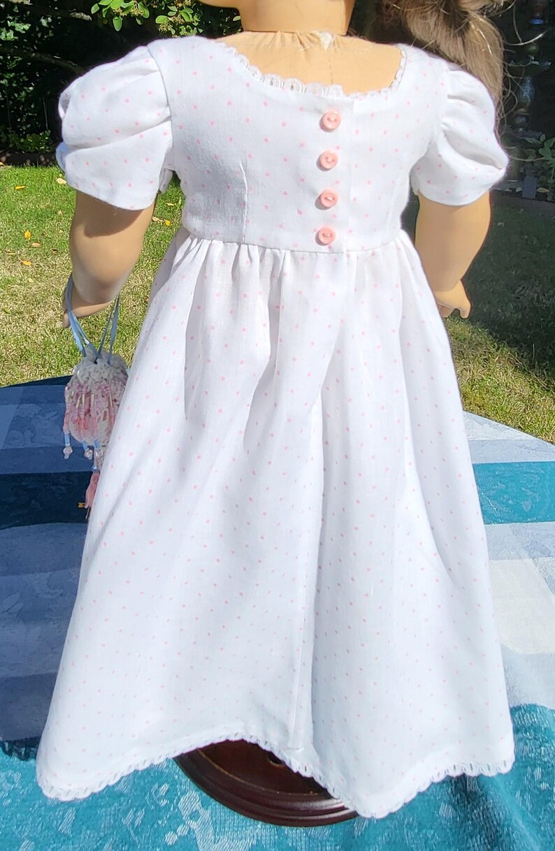 May include: A white dress with small pink polka dots, featuring short puff sleeves, a square neckline with delicate lace trim, and a vertical row of four small pink buttons down the centre of the bodice, paired with a small beaded drawstring bag.
