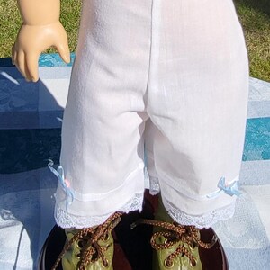 May include: White knee-length doll bloomers featuring a simple waistband, delicate white lace trim at the leg openings, and small light blue ribbon bows on each side, paired with olive green lace-up boots with brown laces.