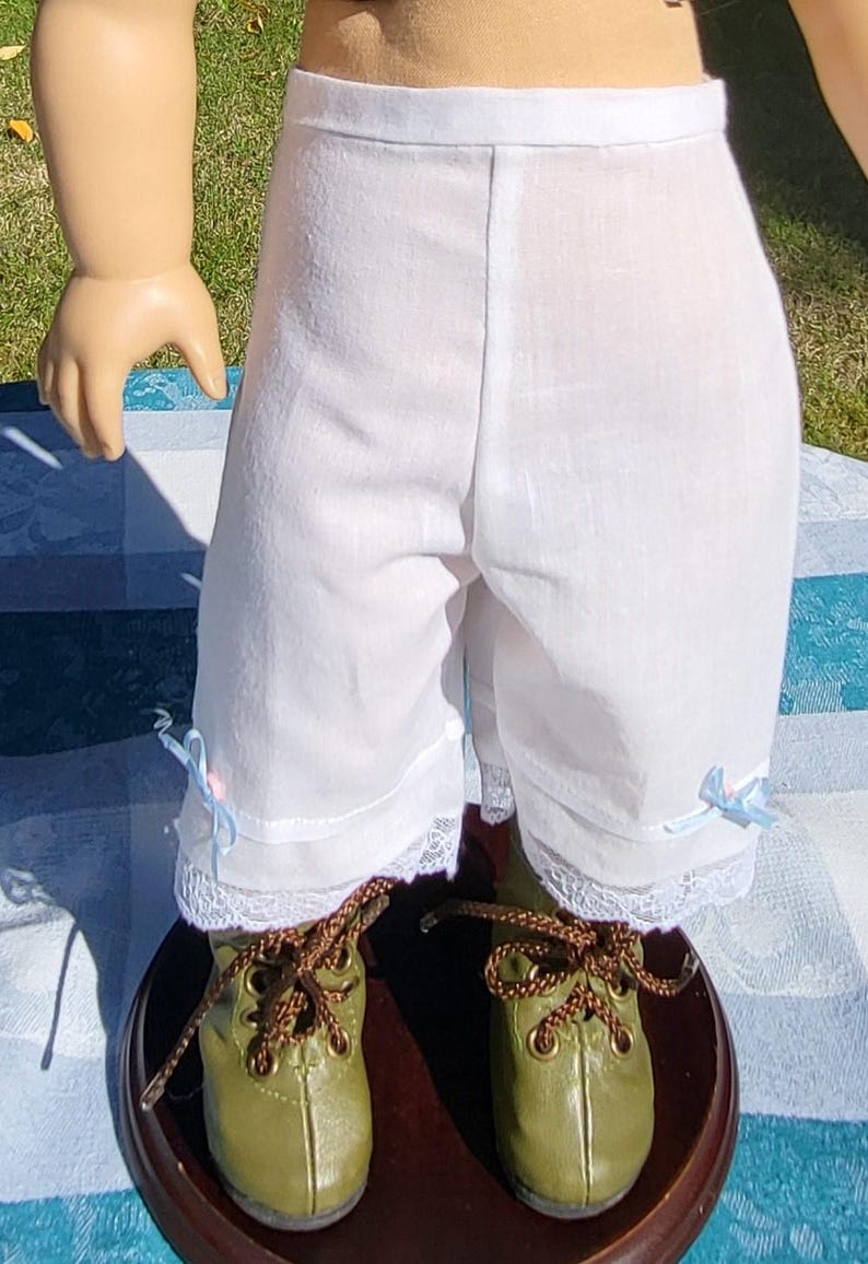 May include: White knee-length doll bloomers featuring a simple waistband, delicate white lace trim at the leg openings, and small light blue ribbon bows on each side, paired with olive green lace-up boots with brown laces.