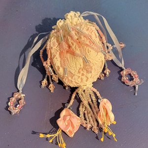 May include: A small, round, cream-coloured lace drawstring pouch adorned with dangling pink glass beads, clear cylindrical beads, and two fabric flower accents with yellow stamens. The pouch features light-coloured ribbon ties with beaded ends.