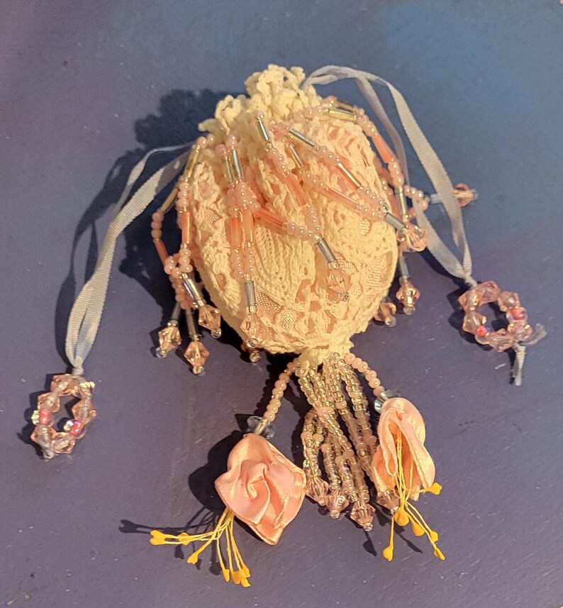 May include: A small, round, cream-coloured lace drawstring pouch adorned with dangling pink glass beads, clear cylindrical beads, and two fabric flower accents with yellow stamens. The pouch features light-coloured ribbon ties with beaded ends.