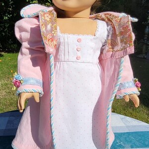 May include: A doll with brown hair wearing a white dress with small pink polka dots and three pink buttons on the bodice, paired with a light pink coat featuring blue and white twisted trim, floral accents on the cuffs, and a patterned lining.
