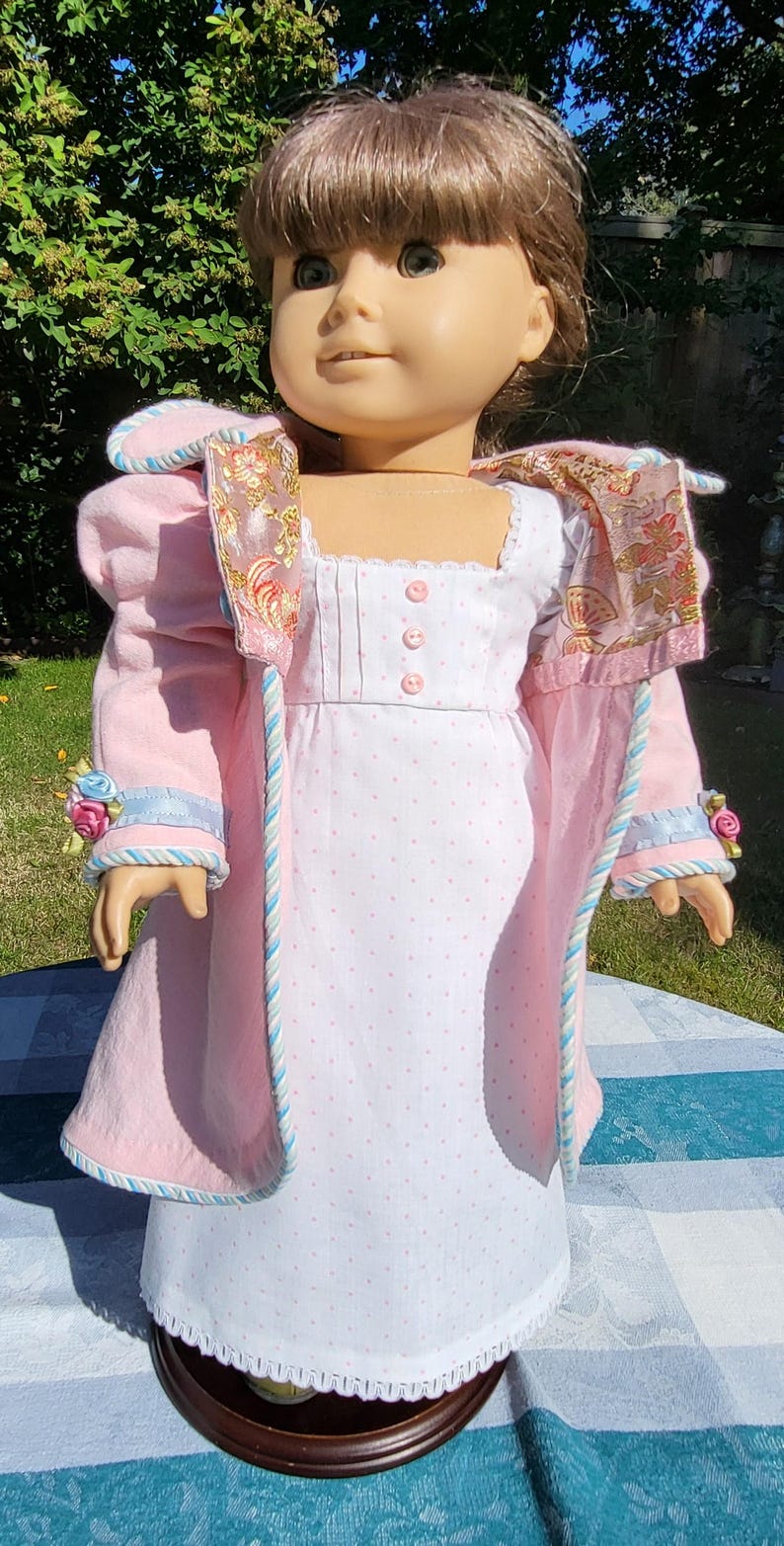 May include: A doll with brown hair wearing a white dress with small pink polka dots and three pink buttons on the bodice, paired with a light pink coat featuring blue and white twisted trim, floral accents on the cuffs, and a patterned lining.