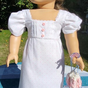 May include: A doll with brown hair wearing a white dress featuring a pink polka dot pattern, puff sleeves, a square neckline, and three pink buttons on the bodice. The doll wears a beaded bracelet and holds a small, decorative drawstring pouch.