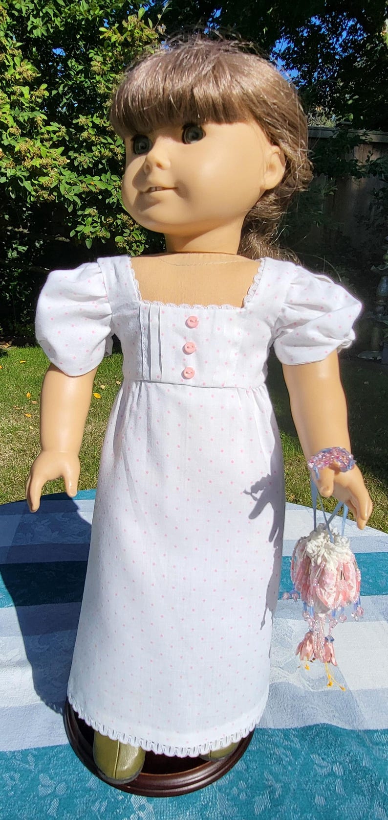 May include: A doll with brown hair wearing a white dress featuring a pink polka dot pattern, puff sleeves, a square neckline, and three pink buttons on the bodice. The doll wears a beaded bracelet and holds a small, decorative drawstring pouch.