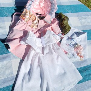 May include: A collection of doll clothing including a pink bonnet with blue lace trim, a pink coat with a floral collar, a white dress with tiny blue dots and lace trim, a white skirt with floral embroidery, and a single olive green lace-up boot.
