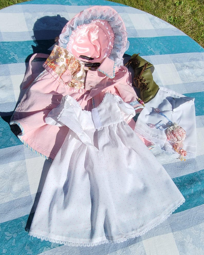 May include: A collection of doll clothing including a pink bonnet with blue lace trim, a pink coat with a floral collar, a white dress with tiny blue dots and lace trim, a white skirt with floral embroidery, and a single olive green lace-up boot.