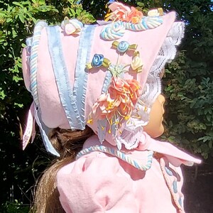 May include: A light pink bonnet featuring light blue ribbon trim, white lace edging, and decorative fabric flowers in shades of peach, cream, and blue. The bonnet is adorned with twisted cord accents and is worn over long brown hair.