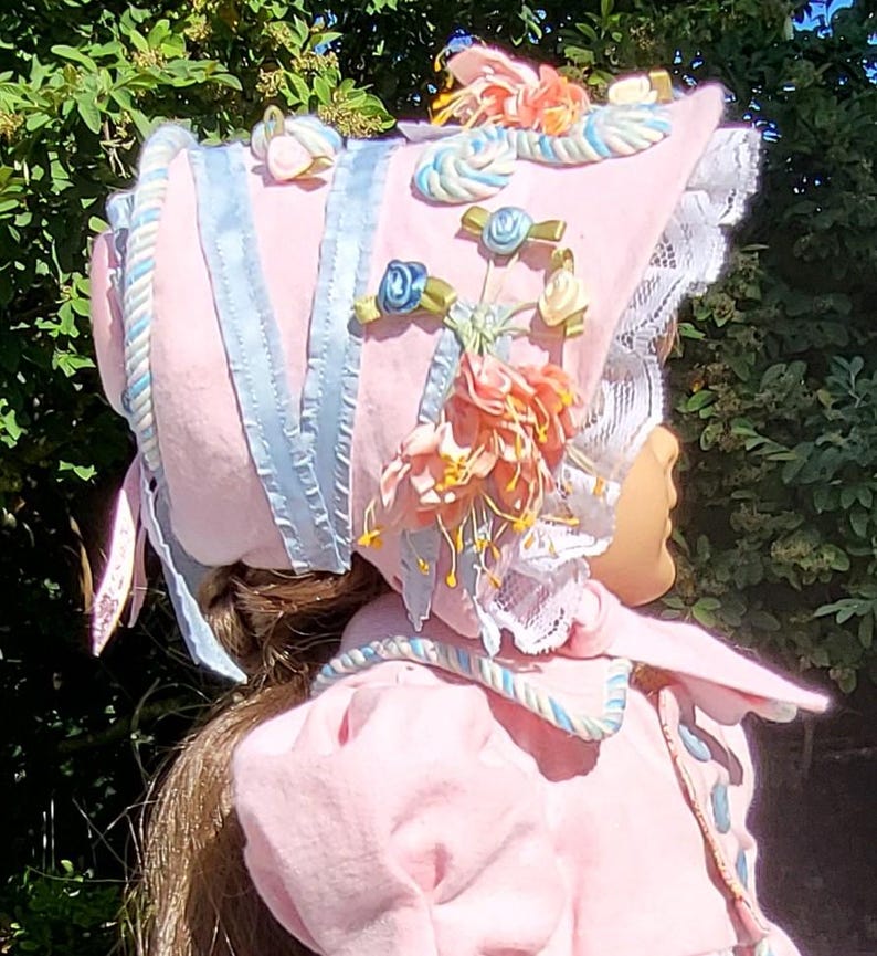 May include: A light pink bonnet featuring light blue ribbon trim, white lace edging, and decorative fabric flowers in shades of peach, cream, and blue. The bonnet is adorned with twisted cord accents and is worn over long brown hair.