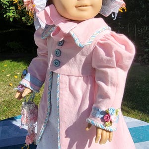 May include: A doll with brown hair wearing a pink coat with blue buttons, a white dress with small dots, and a matching pink bonnet with blue lace trim. The doll features olive green shoes and a small pink floral accessory held in one hand.
