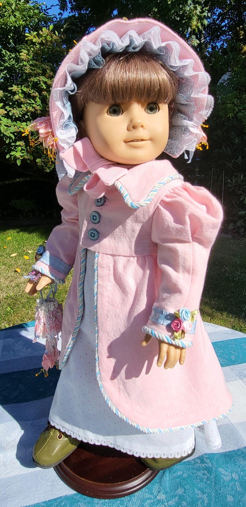 May include: A doll with brown hair wearing a pink coat with blue buttons, a white dress with small dots, and a matching pink bonnet with blue lace trim. The doll features olive green shoes and a small pink floral accessory held in one hand.