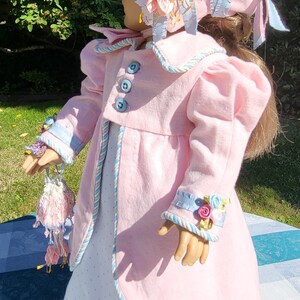 May include: A doll wearing a light pink coat with blue buttons and light blue trim, a white dress with small dots, and a matching pink bonnet decorated with blue and pink ribbon flowers. The doll holds a small, matching pink and white drawstring bag.
