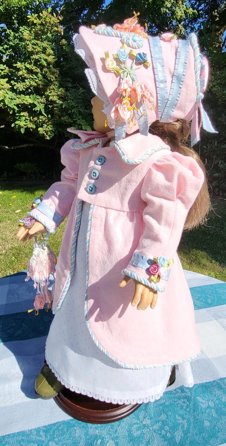 May include: A doll wearing a light pink coat with blue buttons and light blue trim, a white dress with small dots, and a matching pink bonnet decorated with blue and pink ribbon flowers. The doll holds a small, matching pink and white drawstring bag.