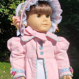 May include: A doll with brown hair wearing a pink coat with blue buttons and blue and white trim. The doll wears a matching pink bonnet with blue lace trim and small floral accents. The coat sleeves feature blue ribbon and small pink and blue fabric flowers.