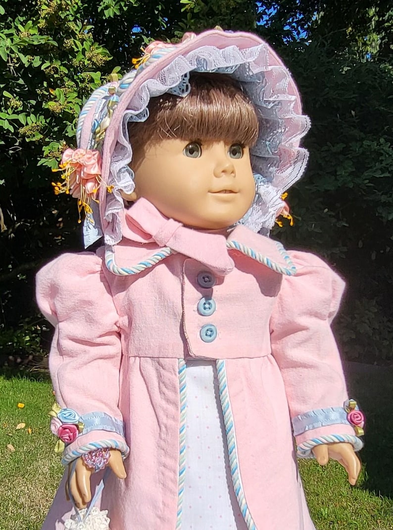 May include: A doll with brown hair wearing a pink coat with blue buttons and blue and white trim. The doll wears a matching pink bonnet with blue lace trim and small floral accents. The coat sleeves feature blue ribbon and small pink and blue fabric flowers.