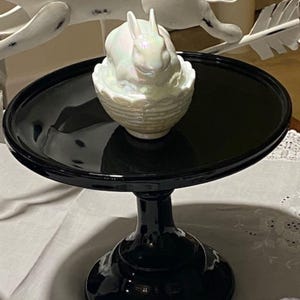 May include: A black, glossy cake stand with a round top and a pedestal base. A pearlescent white bunny figurine sits atop the stand, nestled in a basket-like container. The stand is on a white tablecloth.