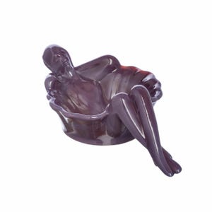 May include: A glossy, dark purple ceramic bowl shaped like a reclining figure. The figure's head and torso form the back of the bowl, while the legs extend over the front edge, creating a sculptural, functional vessel with a smooth, reflective finish.