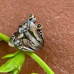 May include: A silver ring with a floral design featuring green gemstones and intricate details. The ring is set on a green plant stem.
