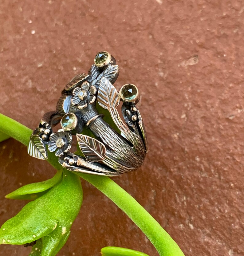 May include: A silver ring with a floral design featuring green gemstones and intricate details. The ring is set on a green plant stem.