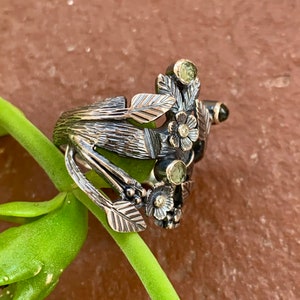 May include: A silver ring with a floral design featuring green and white gemstones. The ring is made of oxidised silver and has a textured finish.
