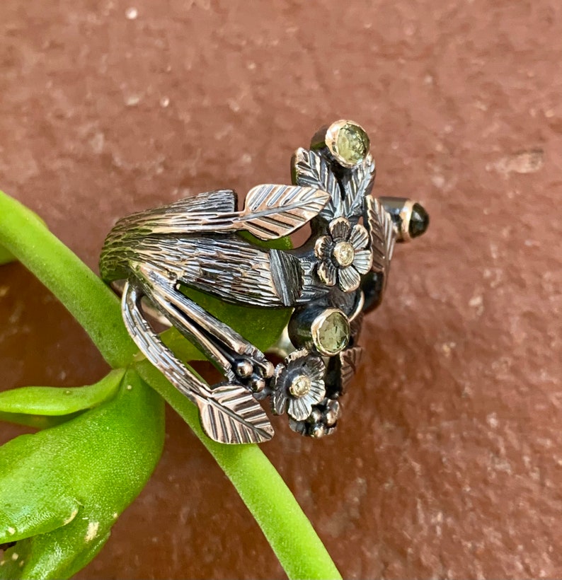 May include: A silver ring with a floral design featuring green and white gemstones. The ring is made of oxidised silver and has a textured finish.