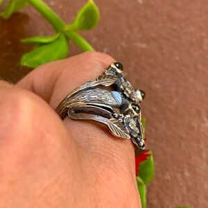 May include: A silver ring featuring a floral design and three green gemstones. The ring is worn on a finger.