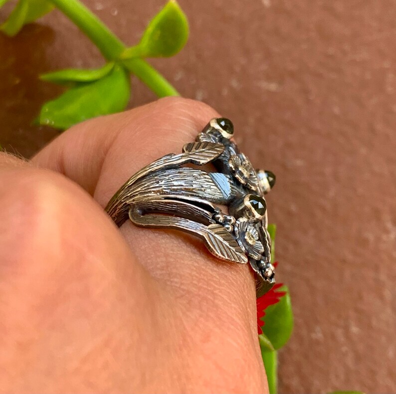 May include: A silver ring featuring a floral design and three green gemstones. The ring is worn on a finger.