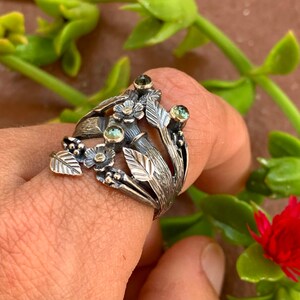 May include: A silver ring featuring a floral design and green gemstones. The ring is worn on a finger, with a blurred background.