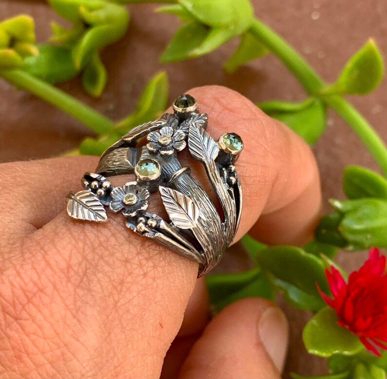 May include: A silver ring featuring a floral design and green gemstones. The ring is worn on a finger, with a blurred background.
