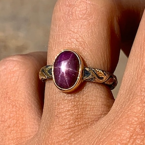 Op de afbeelding: Een gouden ring met een donkerrode ovale robijn-edelsteen, gezet in een bezel-setting. De band is versierd met een gedraaid draadontwerp.