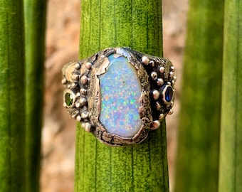 May include: A silver ring featuring a large white opal gemstone set in a textured band. The ring is accented with smaller green and blue gemstones.