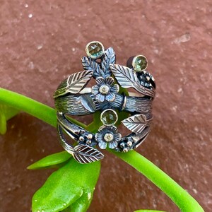 May include: A silver ring with a floral design featuring green and white gemstones. The ring is made of oxidised silver and has a textured finish.