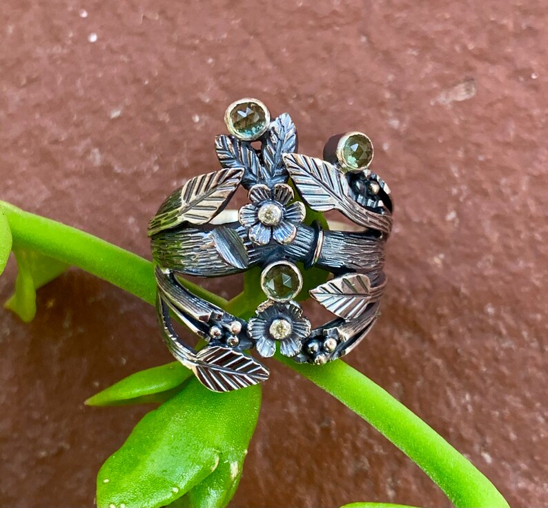 May include: A silver ring with a floral design featuring green and white gemstones. The ring is made of oxidised silver and has a textured finish.