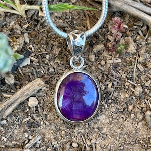 Puede incluir: Un collar con colgante de plata con una gema ovalada de color púrpura intenso. La gema está engastada en un bisel de plata y cuelga de una cadena de plata. El colgante tiene un diseño decorativo y una pequeña piedra transparente.