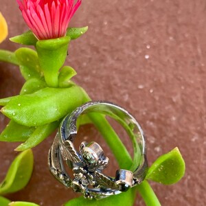 May include: A silver ring with a floral design featuring a green and black gemstone. The ring is set against a backdrop of green foliage and a pink flower.