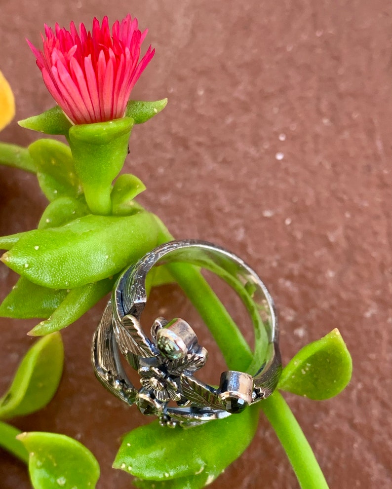 May include: A silver ring with a floral design featuring a green and black gemstone. The ring is set against a backdrop of green foliage and a pink flower.