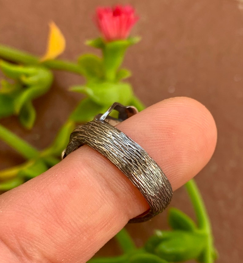 May include: A silver ring with a textured band. The ring is worn on a finger.