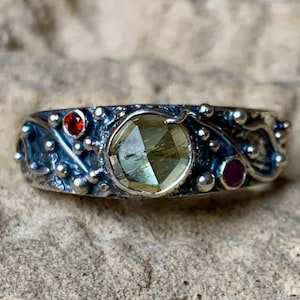 May include: A sterling silver ring with a large, faceted, light green gemstone as the focal point. The ring is adorned with smaller red and pink gemstones and decorative silver accents. The band has a textured, organic design.