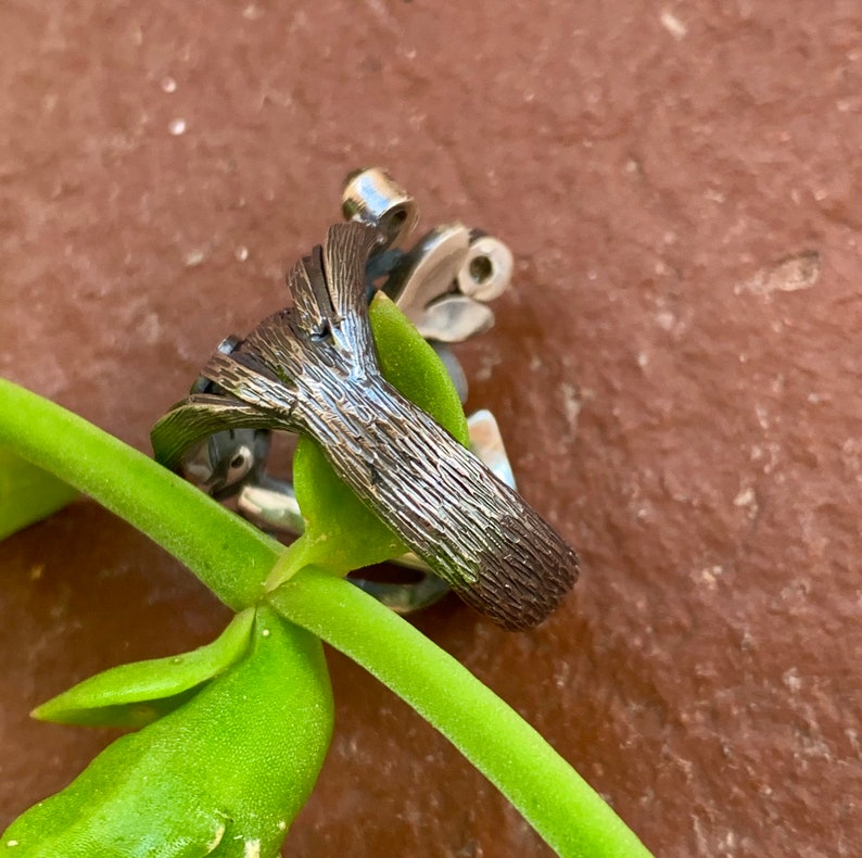 May include: A silver ring featuring a textured tree design. The ring is resting on a green plant stem.