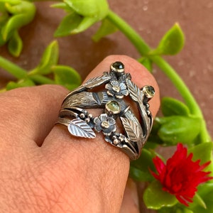 May include: A silver ring with a floral design featuring green gemstones and delicate leaves. The ring is worn on a finger.