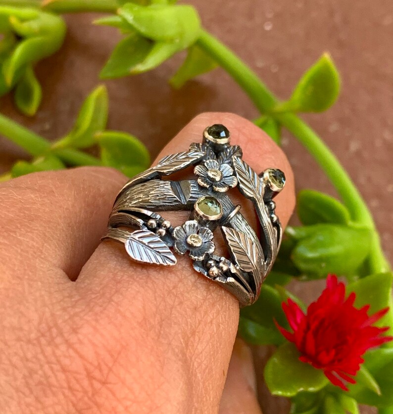 May include: A silver ring with a floral design featuring green gemstones and delicate leaves. The ring is worn on a finger.