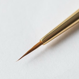 Może przedstawiać: Zbliżenie na mały, cienki pędzel z złotym uchwytem i brązowymi włosiem. Pędzel leży na białej powierzchni.