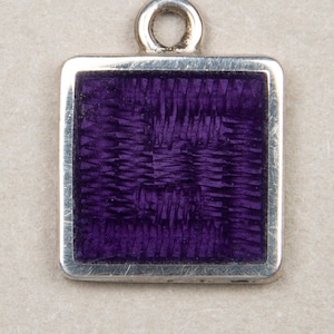 以下が含まれることがあります： 正方形の紫色のエナメルインサートが付いたシルバーチャーム。エナメルは織り模様になっています。