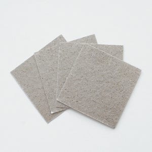 Puede incluir: Cuatro posavasos cuadrados de color gris claro con una textura moteada están abanicados sobre una superficie blanca. Los posavasos están hechos de un material grueso y absorbente, ideal para proteger las superficies de las marcas de agua y los derrames. El color neutro complementa varios estilos de decoración.