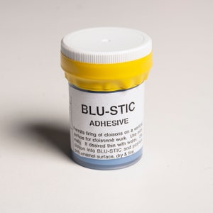 Può includere: Un piccolo barattolo bianco con un coperchio giallo etichettato "BLU-STIC ADHESIVE". L'etichetta include anche istruzioni per l'uso dell'adesivo per lavori di cloisonné.