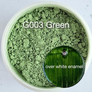 May include: A round container filled with light sage green powder pigment labeled G003 Green. Resting on the powder is a circular dark forest green test tile with a hole at the top, featuring the text over white enamel printed on its surface.