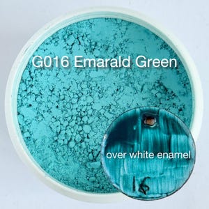 May include: A container of G016 Emerald Green powder pigment with a textured, turquoise appearance. A small, circular, dark teal test tile with a hole at the top is placed on the powder, featuring the text "over white enamel" and "16".