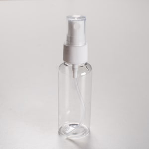 Könnte beinhalten: Eine transparente, leere Sprühflasche mit weißem Sprühkopf. Die Flasche ist zylindrisch und aus transparentem Kunststoff. Die Düse ist weiß mit gerippter Textur, und ein klarer Schlauch erstreckt sich in die Flasche.