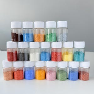 Könnte beinhalten: Eine Sammlung kleiner, klarer Plastikgläser, gefüllt mit farbigen Pulvern. Die Pulver sind in verschiedenen Farbtönen, darunter Rot, Blau, Orange, Grün und Gelb. Jedes Glas hat einen weißen Schraubverschluss. Die Gläser sind in einer ordentlichen, gestapelten Anordnung angeordnet.