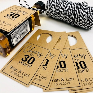 May include: Brown paper bottle tags with a black circle hole at the top. The tags have black text that reads "Cheers to 30 years!" with the names "Brian & Lori" and the date "10.29.2019".